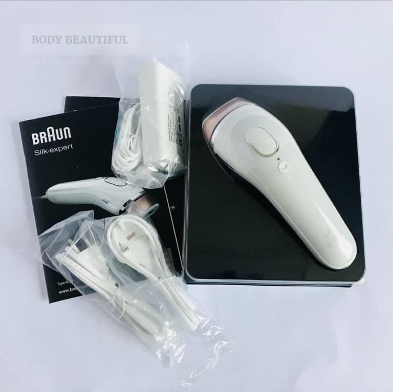 The contents of the Braun Silk expert box laid on a white surface.: x2 user manuals to cover European languages, the Silk-expert IPL device in protective moulded, clear plastic, the power pack and a UK and European socket power cable. They're all neatly contained in small plastic bags with cable ties. 